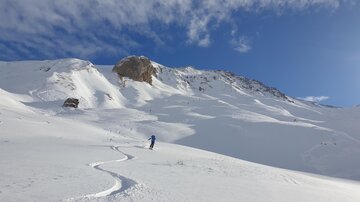 A snowy mountain landscape with a clear blue sky. In the foreground, a person is making tracks in the snow.