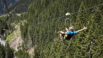 A person glides on a zip line over a forest of tall fir trees. The landscape is green and mountainous, ideal for adventures in nature.