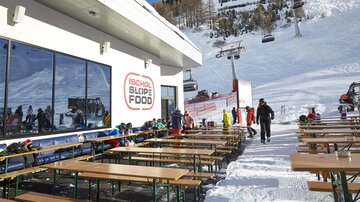 Ein modernes Almhüttencafé mit Sitzplätzen im Freien, umgeben von schneebedeckten Bergen. Skifahrer und snowboarder genießen den Tag im Winterparadies.