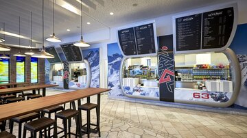 Ein modernes Café mit hellen Holzstühlen und großen Fenstern. An der Wand sind Werbetafeln und eine ansprechende Theke mit bunten Getränken und Snacks zu sehen.