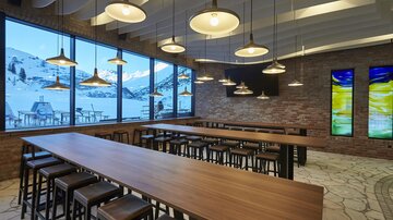 Ein modernes Restaurant mit großen Fenstern und Blick auf die schneebedeckten Berge. Der Raum ist hell erleuchtet und verfügt über lange Holztische und Stühle.