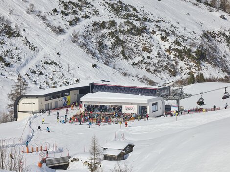 A bustling ski resort in the mountains with many skiers. In the background, snow-covered slopes and a cable car can be seen.