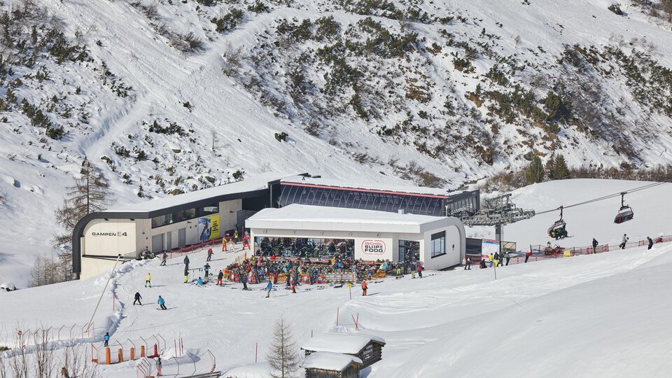 Eine belebte Skistation in den Bergen mit vielen Skifahrern. Im Hintergrund sind schneebedeckte Hänge und eine Seilbahn zu sehen.