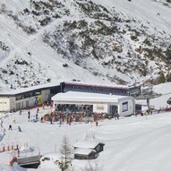 Eine belebte Skistation in den Bergen mit vielen Skifahrern. Im Hintergrund sind schneebedeckte Hänge und eine Seilbahn zu sehen.