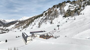 Eine malerische Schneelandschaft mit einem Skilift und einer Berghütte. Im Hintergrund sind schneebedeckte Berge und ein blauer Himmel zu sehen.