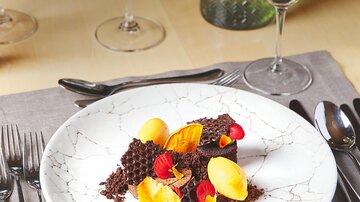 An elegantly arranged plate with dark chocolate, yellow fruit, and edible flowers. In the background, wine glasses and green decoration are visible.