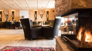 A cozy restaurant with elegant armchairs and an open fireplace. In the background, guests can be seen eating and chatting.