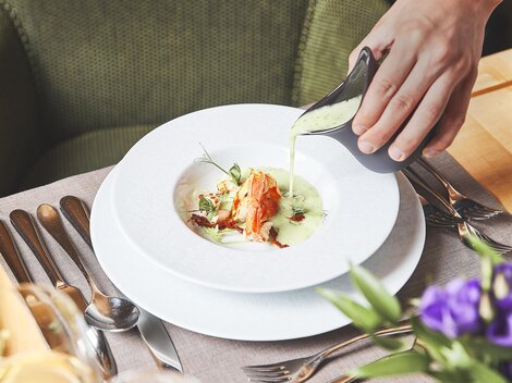 An elegant dish is served on a white plate. A hand pours a green sauce over the food, which is garnished with herbs.