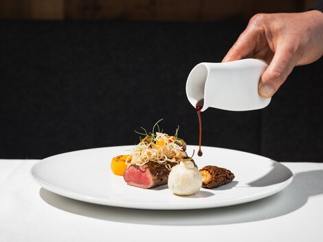 Eine stilvolle Speisepräsentation auf einem weißen Teller. Eine Hand gießt eine Sauce über das Gericht, das aus Fleisch, buntem Gemüse und Beilagen besteht.