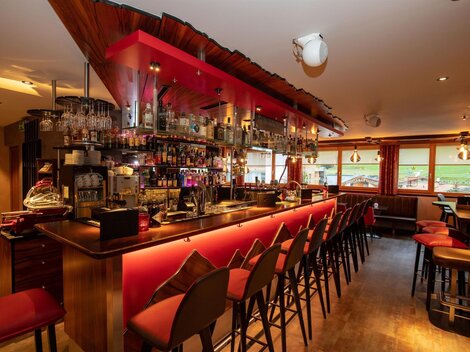 A modern bar with an inviting use of red light. The counter is well-stocked and there are many chairs waiting for guests.