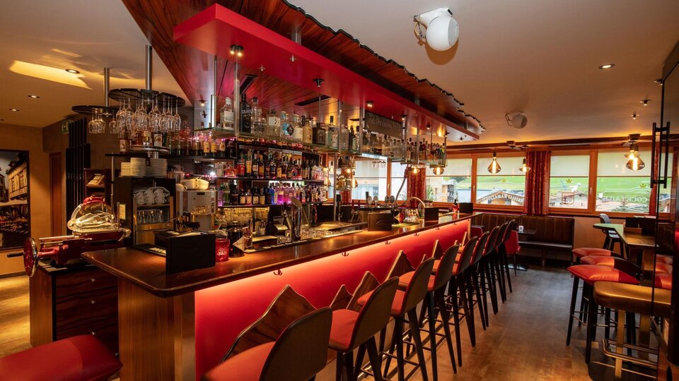 A modern bar with an inviting use of red light. The counter is well-stocked and there are many chairs waiting for guests.