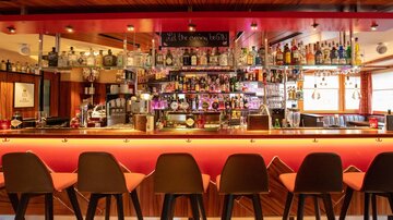 A stylish bar with a long counter and modern bar stools. In the background, many alcoholic beverages are displayed on a shelf.