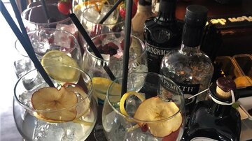 A selection of cocktails in large glasses with fresh fruits and herbs. In the background, various bottles of spirits are visible.