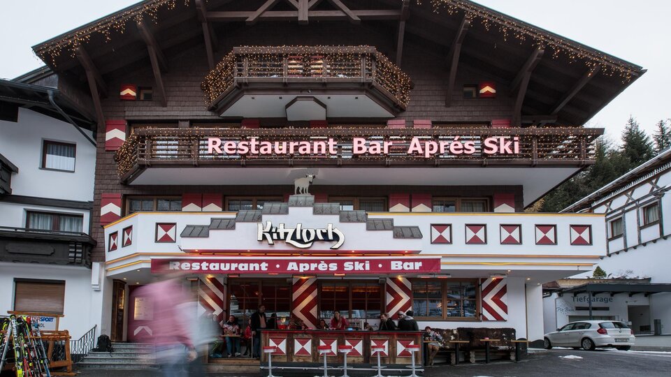 A cozy restaurant with an après-ski style bar. The façade is rustic with wooden details and an inviting outdoor area.