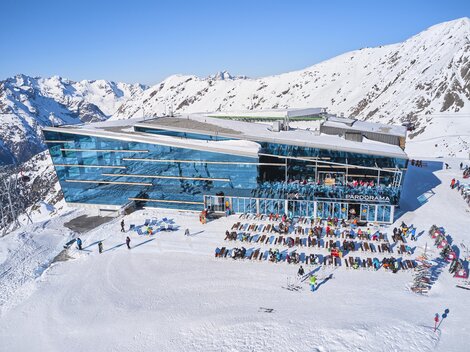 A modern mountain cabin surrounded by snow-covered mountains. Many people are enjoying the sunny day outdoors.
