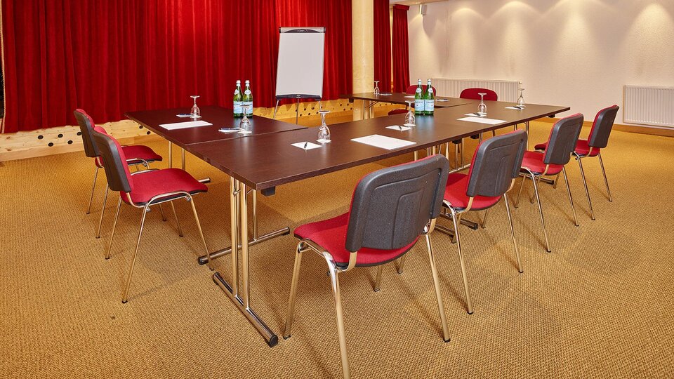 A modernly equipped meeting room with a large table and red chairs. The walls are decorated with red curtains and soft lighting.
