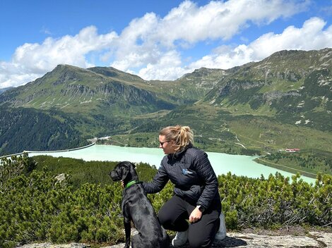 A woman sits with her dog on a rock and gazes at a green landscape with mountains and a lake. The sky is blue with some clouds.