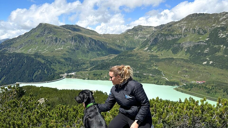 A woman sits with her dog on a rock and gazes at a green landscape with mountains and a lake. The sky is blue with some clouds.