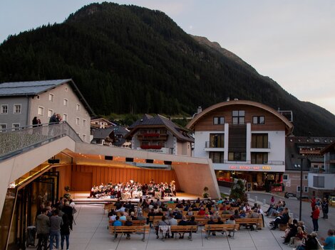 An outdoor event with an orchestra is playing against a backdrop of mountains. Many people are sitting on benches and enjoying the music.
