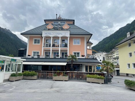 A charming hotel with a bright facade and a balcony on the ground floor. In the background, green mountains and a tranquil atmosphere can be seen.