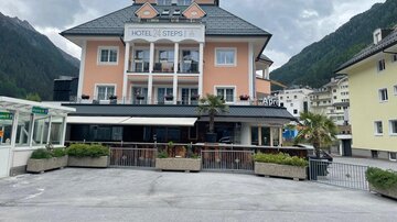 Ein charmantes Hotel mit einer hellen Fassade und Balkon im Erdgeschoss. Im Hintergrund sind grüne Berge und eine ruhige Atmosphäre zu sehen.