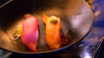 Two pieces of sushi on a black plate. One piece is pink and the other is orange, garnished with herbs.