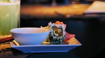 A plate with sushi and a small bowl of sauce. In the background, a drink is visible.