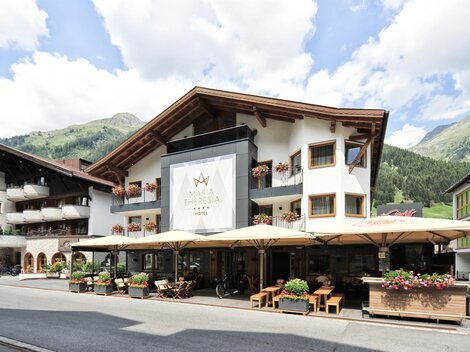 A cozy hotel with an inviting outdoor area and terraces. In the background, green mountains and a blue sky can be seen.