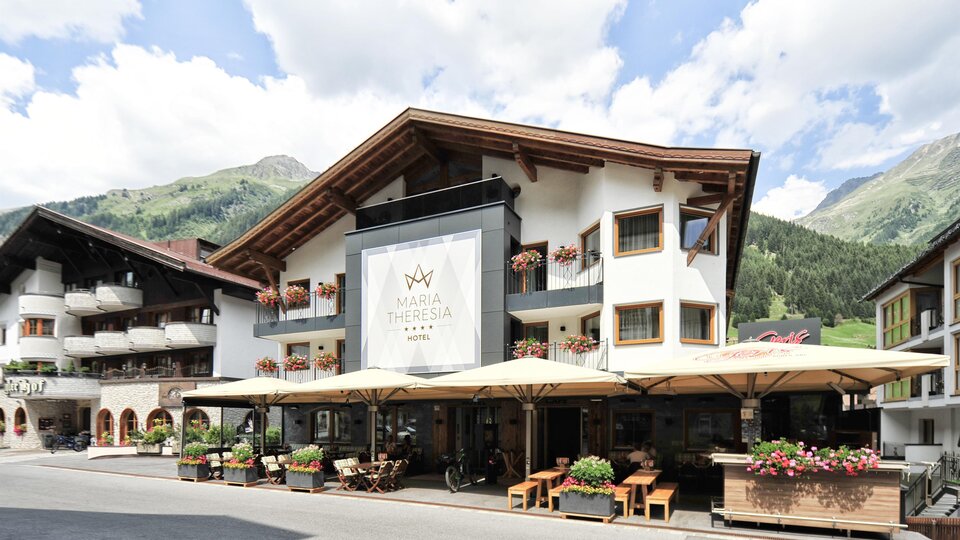 Ein gemütliches Hotel mit einem einladenden Außenbereich und Terrassen. Im Hintergrund sind grüne Berge und ein blauer Himmel zu sehen.