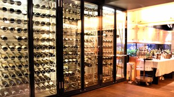 A stylish wine cellar with large glass shelves showcasing a variety of wines. In the background, there is a bar with various drinks visible.