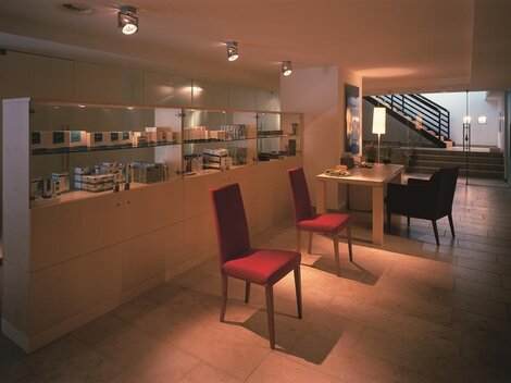 A bright reception area with modern furniture and shelves displaying care products. Two red chairs are placed in front of a table, while a staircase is visible in the background.