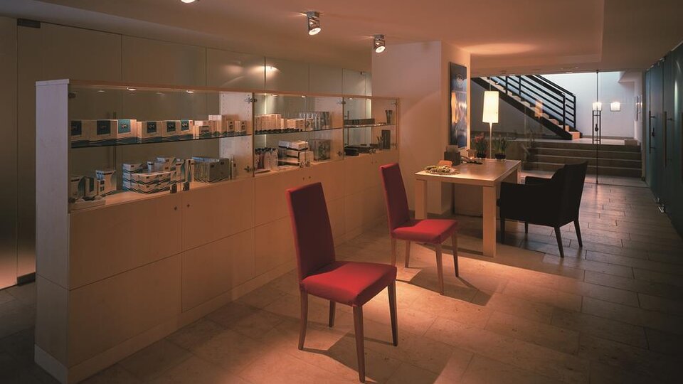 A bright reception area with modern furniture and shelves displaying care products. Two red chairs are placed in front of a table, while a staircase is visible in the background.