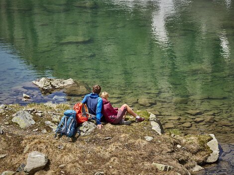 Eine ruhige Landschaft am Ufer eines klaren Sees. Zwei Personen sitzen entspannt auf den Steinen und genießen die Aussicht.