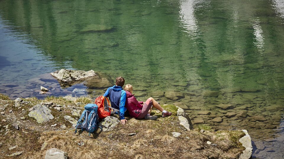 Eine ruhige Landschaft am Ufer eines klaren Sees. Zwei Personen sitzen entspannt auf den Steinen und genießen die Aussicht.