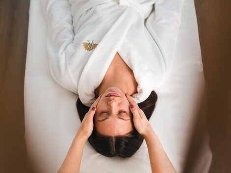 A woman is lying relaxed on a lounger receiving a facial massage. She is wearing a white bathrobe and looks happy.