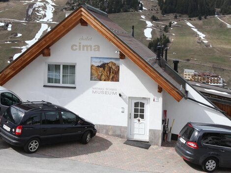 A cozy building with a wooden roof and a sign saying "Museum". Parking for several cars is available, and snow-covered hills are visible in the background.