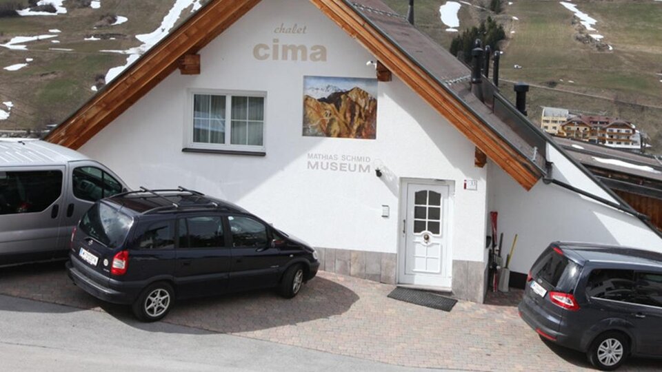 A cozy building with a wooden roof and a sign saying "Museum". Parking for several cars is available, and snow-covered hills are visible in the background.