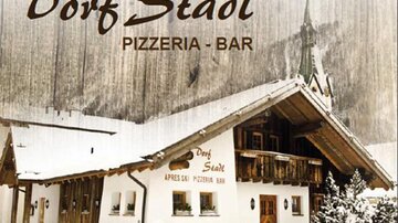 A cozy building in alpine style named "Dorf Stadl". It is a pizzeria and bar, surrounded by snow-covered mountains.
