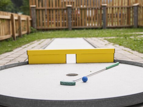 Ein Minigolfplatz mit einer runden Spielfläche und einer gelben Zielwand. Der Ball und der Schläger sind bereit zum Spielen.