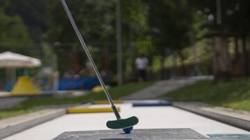 Eine Minigolfbahn mit einem Putter und einer Zielfläche ist im Vordergrund. Im Hintergrund sind Spielgeräte und grüne Wiesen zu sehen.