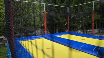 Ein Trampolin in einem Garten, umgeben von Bäumen. Die Sprungfläche ist gelb und die Umzäunung ist rot.
