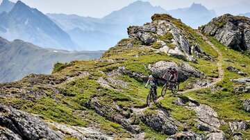 Two mountain bikers ride along a narrow path through a mountainous, green landscape. In the background, impressive mountains and a clear sky can be seen.