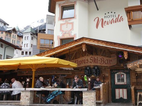 A cozy restaurant with a wooden terrace and a large sunshade. People are sitting together and enjoying drinks while the mountains are visible in the background.