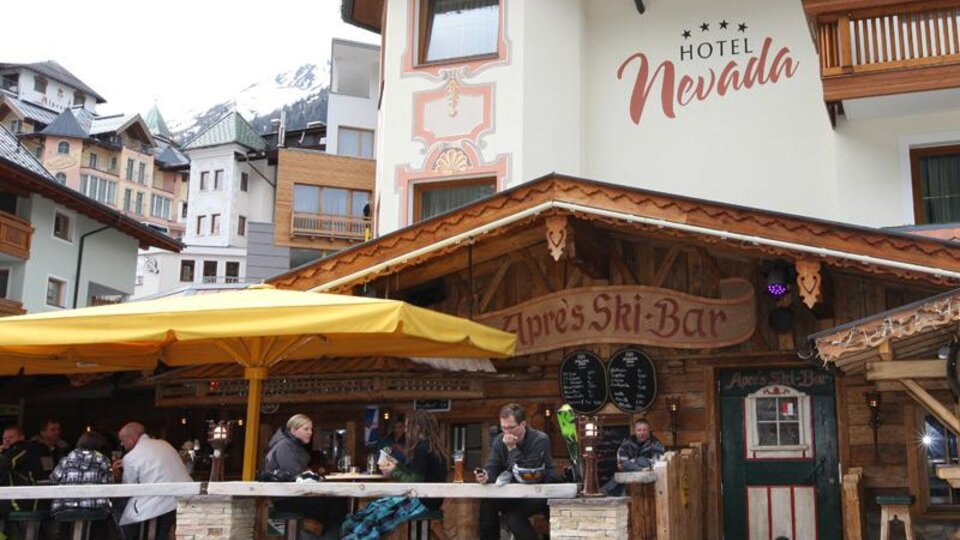 A cozy restaurant with a wooden terrace and a large sunshade. People are sitting together and enjoying drinks while the mountains are visible in the background.