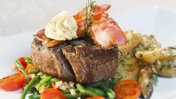 A juicy steak on a bed of vegetables and potatoes, garnished with bacon and herb butter. Fresh tomatoes and green beans complete the dish.