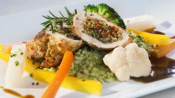 A artistically arranged plate with stuffed chicken, surrounded by colorful vegetables such as carrots, broccoli, and cauliflower. On the plate, there is also green risotto and a tasty sauce.