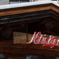 Ein traditionelles Holzgebäude mit einem schneebedeckten Dach. Das Schild mit der Aufschrift "Nikis Stube" ist gut sichtbar.