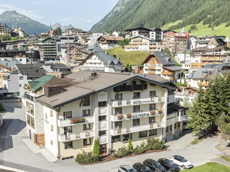 A picturesque alpine village with typical chalets and a beautiful mountain landscape. In the foreground, there is a modern building with parking spaces.
