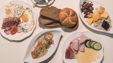 Ein reichhaltiges Frühstück mit Rührei, Speck, frischem Brot und Obst. Die Auswahl umfasst verschiedene Aufschnittsorten und Müsli.