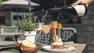 Ein stilvolles Essen im Freien mit einer Flasche Rosé, die in ein Glas gegossen wird. Auf dem Tisch befinden sich auch frisches Brot und eine kleine Pflanze in einem Topf.